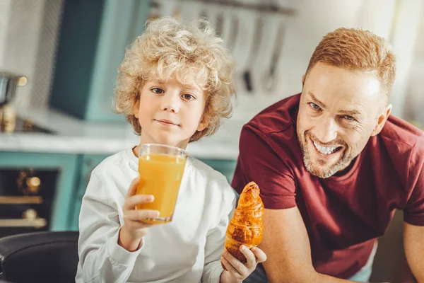 Cute curly boy eating croissant and drinking orange juice — Stock Photo, Image