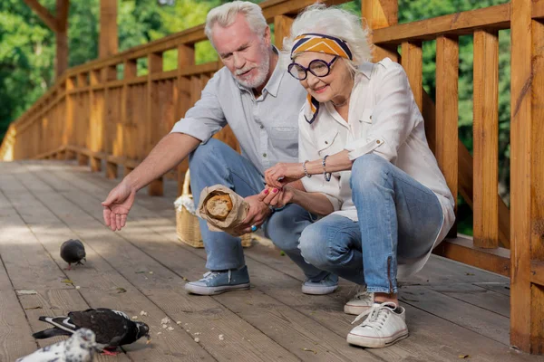 Happy starších lidí sedí na mostě a krmení ptáků s chlebem — Stock fotografie