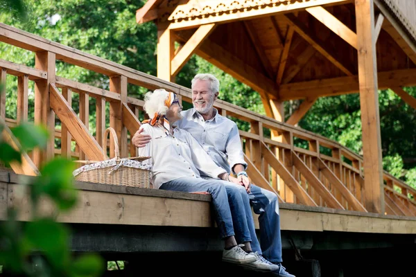 Feliz pensionista abrazando a su esposa mientras está sentado en el puente con ella — Foto de Stock