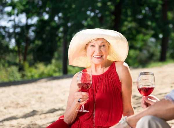 Pozitív nyugdíjas kalapot visel, és mosolyogva, miközben ivott bort — Stock Fotó