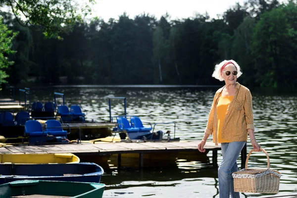 Розслаблена літня жінка тримає кошик і ходить на пірсі — стокове фото