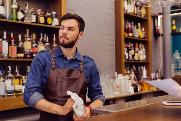 Curioso barman atenciosamente olhando para a distância enquanto a limpeza de óculos — Fotografia de Stock