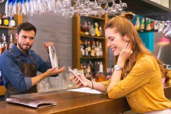 Menina alegre olhando para seu smartphone e o barman fazendo bebidas — Fotografia de Stock