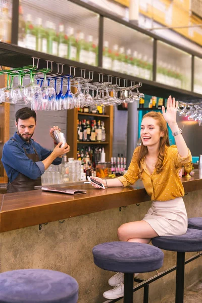 Feliz joven sentada en el mostrador del bar y saludando a su amigo — Foto de Stock