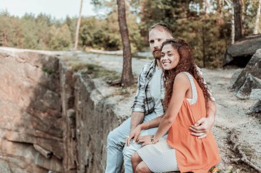 Sarışın adam kayanın üzerinde otururken gülümseyen kız arkadaşı sarılma sakallı