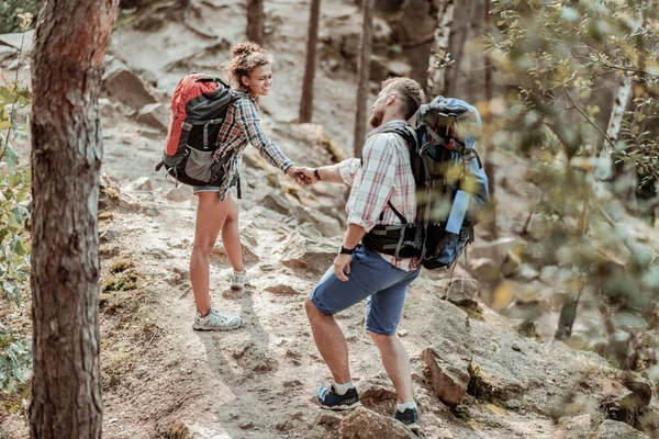 Curly beaming woman with backpack holding hand of her man while hiking — Stock Photo, Image