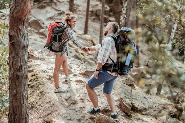 Curly beaming woman with backpack holding hand of her man while hiking
