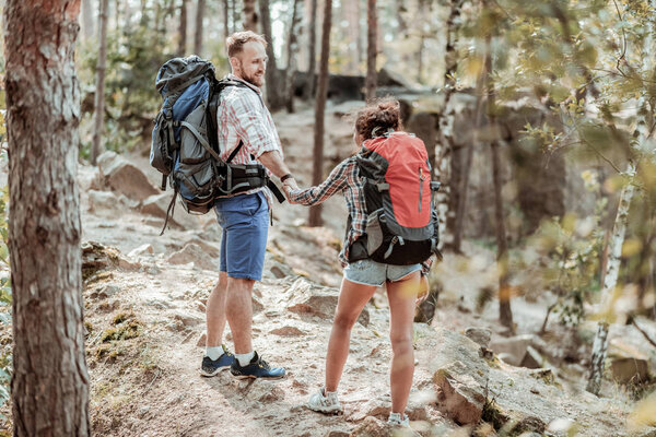 Bearded man with backpack supporting his tender woman while hiking