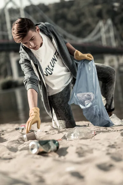 Joven estudiante guapo proporcionando trabajo voluntario mientras recoge basura en la playa — Foto de Stock