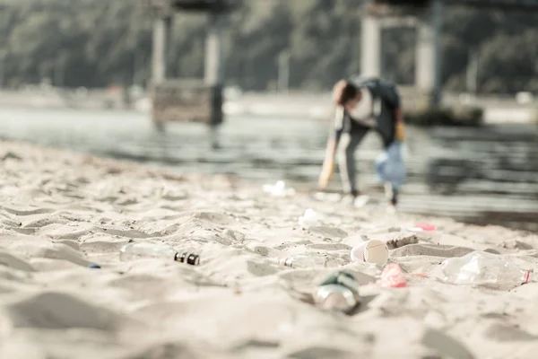 Cerca de la basura que yace en la arena mientras un joven voluntario la recoge —  Fotos de Stock