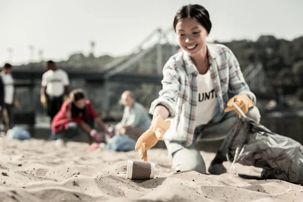 Улыбающаяся социально активная женщина в перчатках собирает пустые кофейные чашки на пляже — стоковое фото