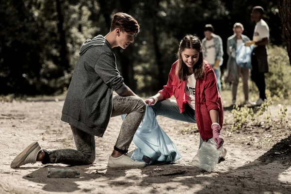 Nuttig dark-haired jongen komen aan zijn vriend en helpen haar reiniging Prullenbak in bos — Stockfoto