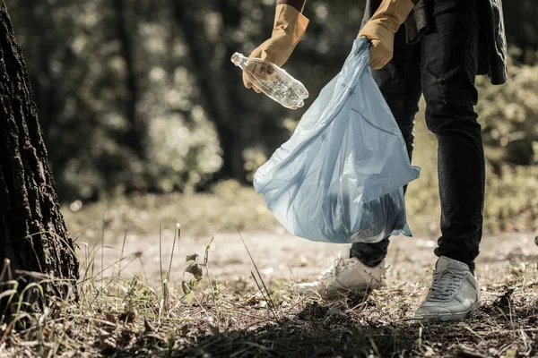 Joven alumno con jeans y zapatillas de deporte recogiendo botellas vacías en el bosque — Foto de Stock