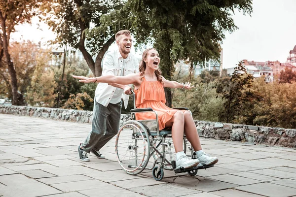 Emocionado chica poniendo manos arriba y novio empujando su silla de ruedas — Foto de Stock