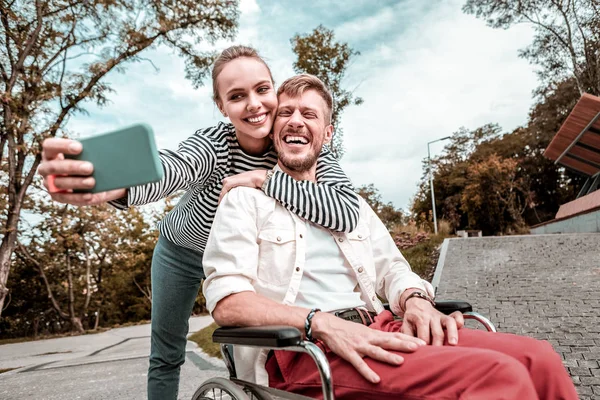 Hombre en silla de ruedas riendo mientras linda novia tomando selfie con él — Foto de Stock