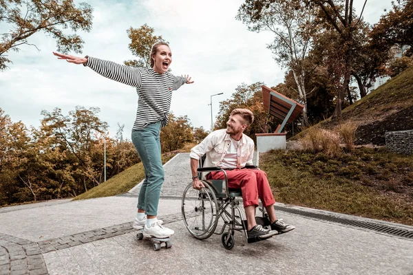 Hombre positivo sentado en silla de ruedas y su novia montando su monopatín — Foto de Stock