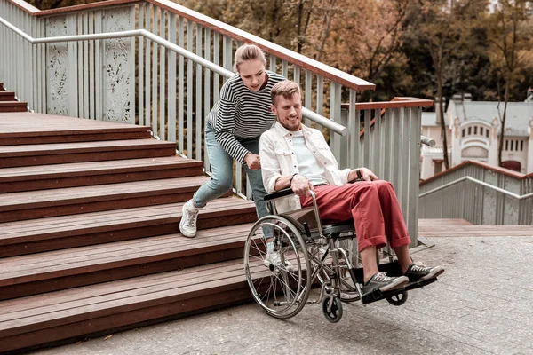 Mujer fuerte ayudando a un hombre inválido y llevando su silla de ruedas — Foto de Stock