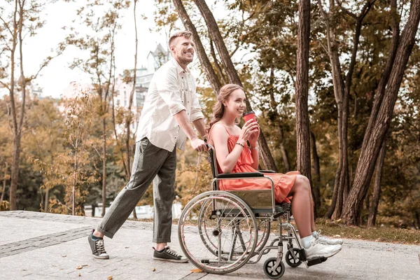 Homem feliz empurrando a cadeira de rodas de sua namorada enquanto caminha no parque — Fotografia de Stock