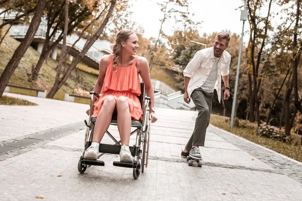 Chica positiva montando su silla de ruedas y novio activo de pie en penny board — Foto de Stock