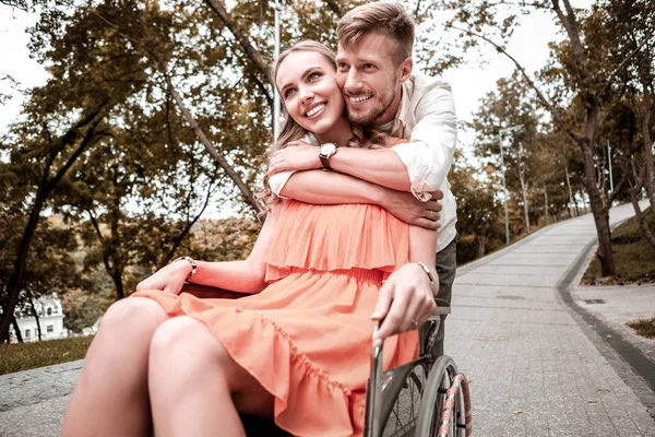 Glad tjej som sitter i rullstolen och hennes pojkvän kramar hennes axlar — Stockfoto