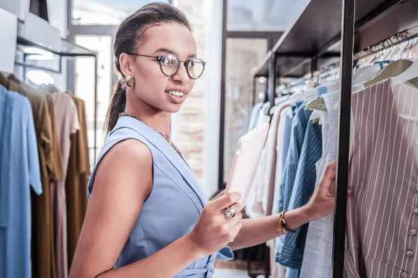 Mujer morena usando gafas sintiéndose alegre eligiendo ropa nueva — Foto de Stock