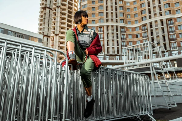 Suntanned lady sitting on the metal fencing taking off her red overcoat — Stock Photo, Image