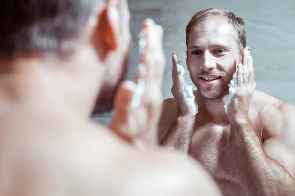 Cheerful blue-eyed man smiling while looking into mirror and shaving face — Stock Photo, Image