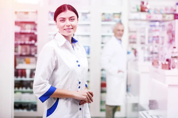 Mulher sorridente vestindo uniforme branco trabalhando como farmacêutica em uma farmácia — Fotografia de Stock