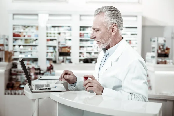 Farmacéutico maduro barbudo en bata blanca trabajando duro usando su portátil de plata — Foto de Stock