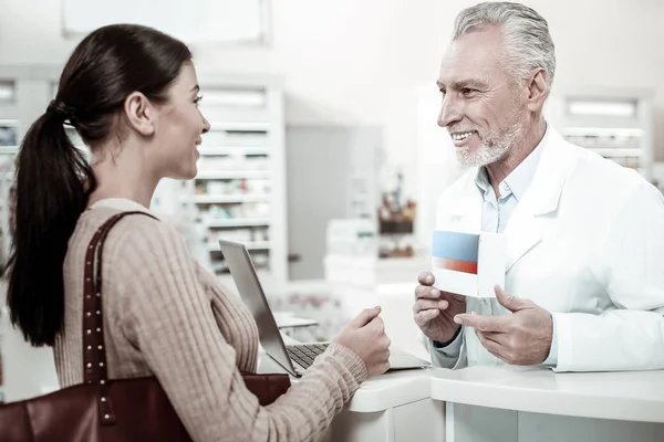 Agradable sonriente farmacéutico barbudo recomendando buenas píldoras a su visitante —  Fotos de Stock