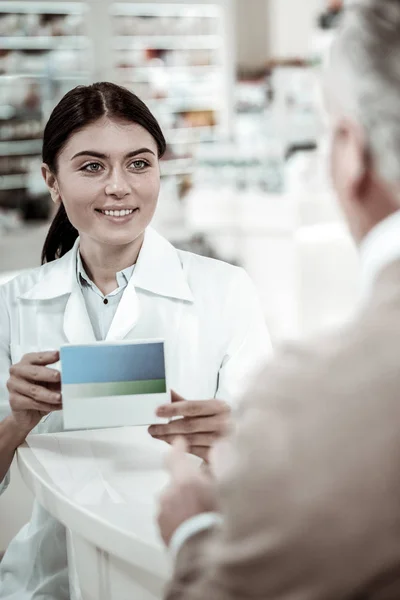 Farmacêutica de cabelos escuros usando casaco branco vendendo pílulas boas seu visitante — Fotografia de Stock