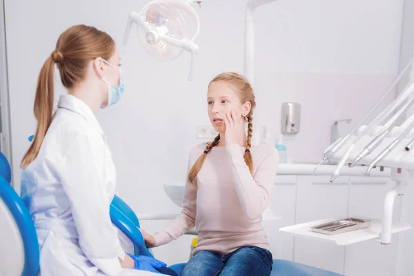 Menina tocando sua bochecha enquanto fala com um dentista pediátrico — Fotografia de Stock