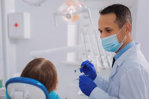 Guapo dentista sosteniendo instrumentos y mirando a su paciente — Foto de Stock
