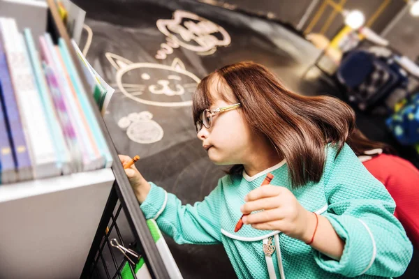 Cute pleasant girl with Down syndrome drawing cute cat on blackboard — Stock Photo, Image