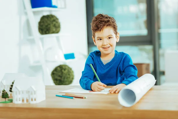 Joyeux garçon assis à la table et souriant tout en dessinant — Photo