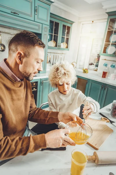 Cute blonde-haired boy holding utility whisk while cooking omelet