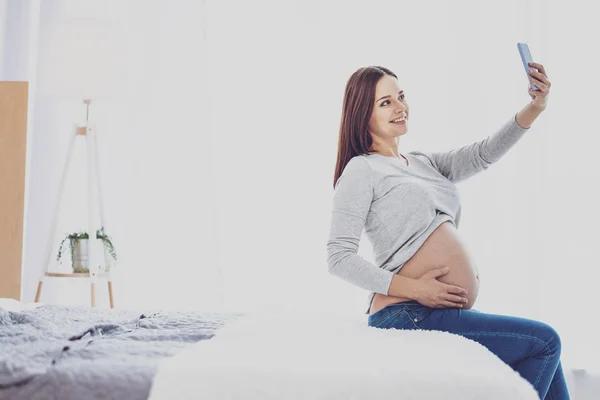 Glad gravid kvinna tar selfie sittande på sängen — Stockfoto