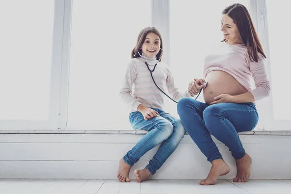 Glad tjej lyssnar på barnet i magen mödrar med stetoskop — Stockfoto