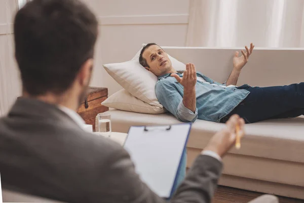 Homem estressado deitado no sofá bege explicando seus problemas ao terapeuta — Fotografia de Stock