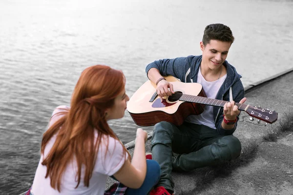 잘생긴 로맨틱 남자 친구 강 근처에 앉아 그의 여자 친구를 위해 기타를 연주 — 스톡 사진