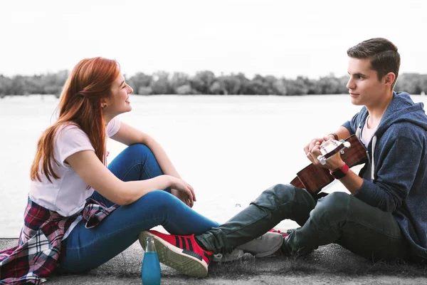 Пара счастливых сияющих подростков наслаждаются прекрасным вечером с гитарой у реки — стоковое фото
