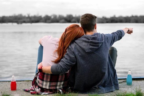 Dark-haired boyfriend hugging his girlfriend sitting near river and drinking alcohol — Stock Photo, Image