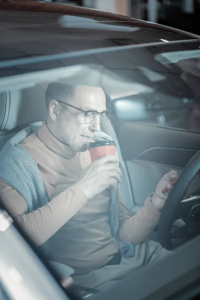 Бизнесмен пьет кофе на вынос и пользуется телефоном, сидя в машине — стоковое фото