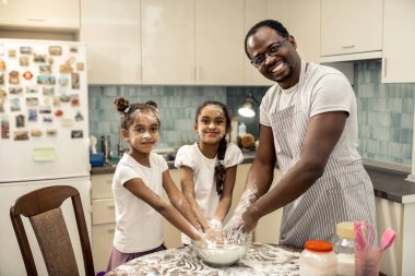 Father in striped apron and two cute daughters cooking pie for mother