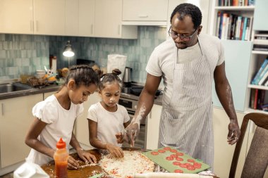 Father wearing striped apron helping his little girls making pizza