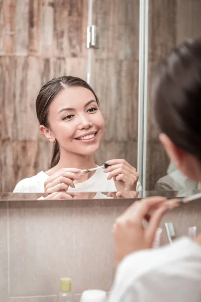 Mujer de ojos oscuros sonriendo mientras se mira en el espejo y haciendo maquillaje — Foto de Stock