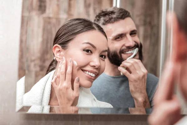 Hermosa mujer usando crema facial mientras el hombre se cepilla los dientes — Foto de Stock