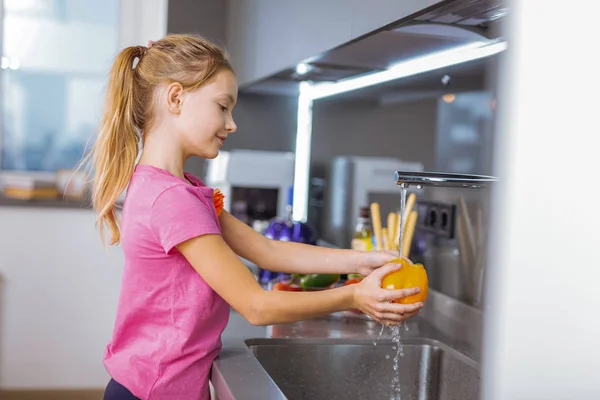 Chica bonita de pelo largo en una camisa rosa lavando verduras en la cocina —  Fotos de Stock