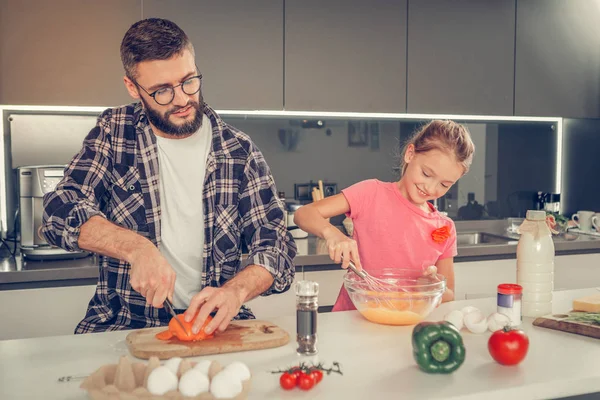 Dulce chica de pelo rubio en una camiseta rosa y su padre cocinando tortilla juntos — Foto de Stock