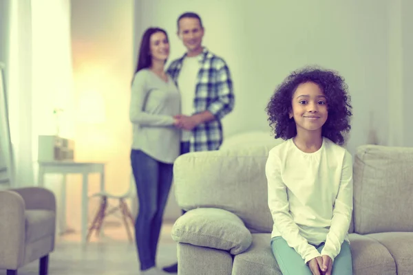 Kleines Mädchen sitzt auf der Couch, ihre Eltern stehen hinter ihr. — Stockfoto